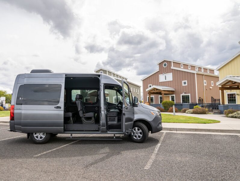 A gray passenger van with the side door open is parked in a lot, ready for a wine tour. The background features winery buildings and a cloudy sky.