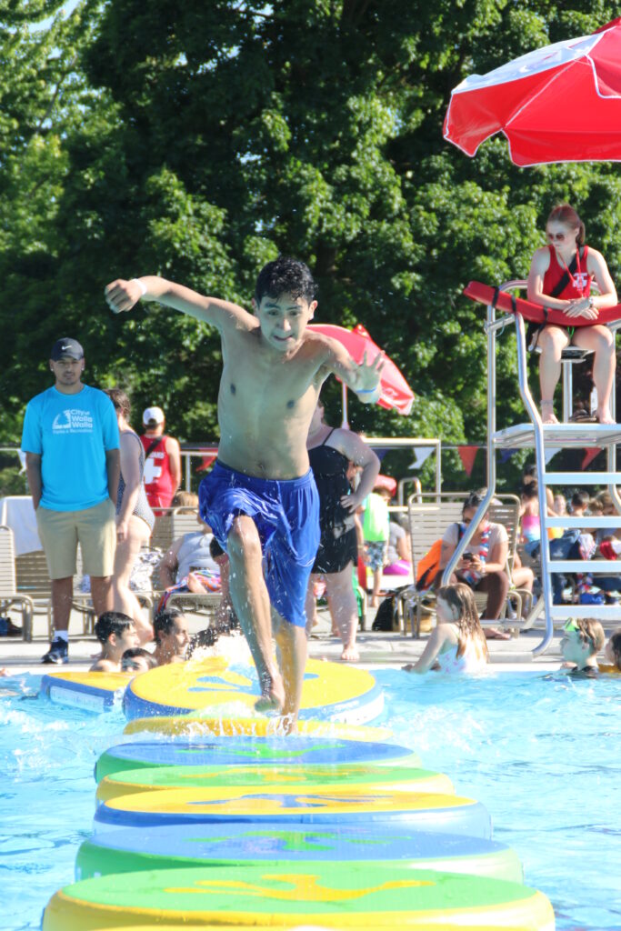 A kid with a gritty face running across floating platforms in the water at the pool. Various pool-goers in the background.